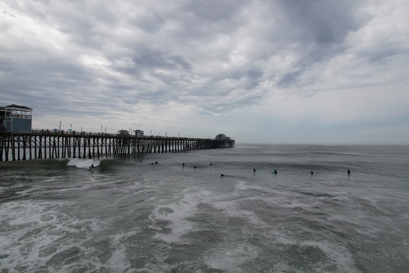 Surfers riding waves next to the Oceanside Pier on an overcast day